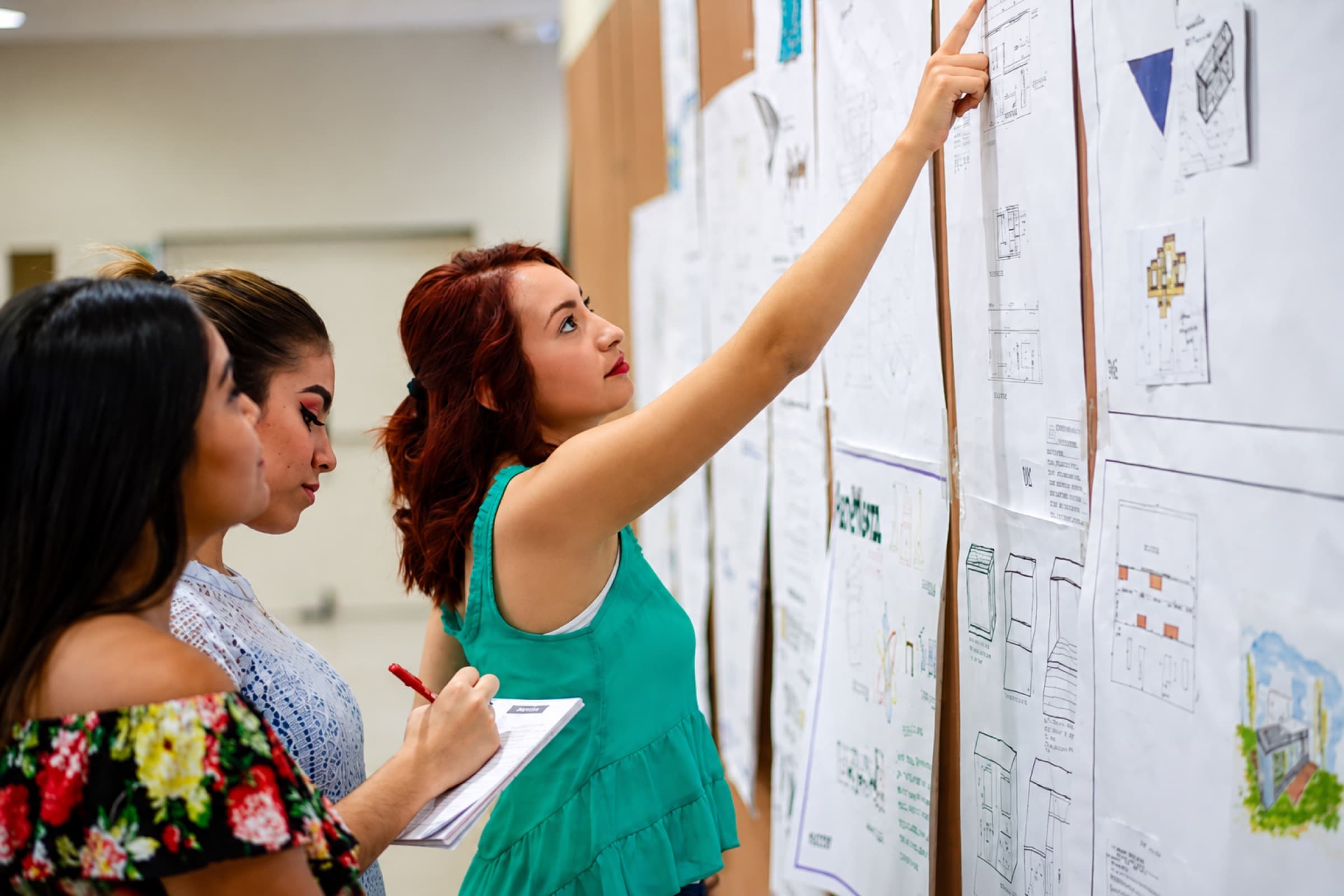 Three colleagues at a wall of pinned plans, one pointing at a sheet while another takes notes